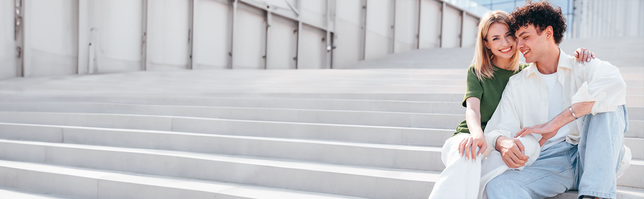 Et ungt par holder on hinanden på en trappe - hun smiler ind i kameraet mens han ser ned.