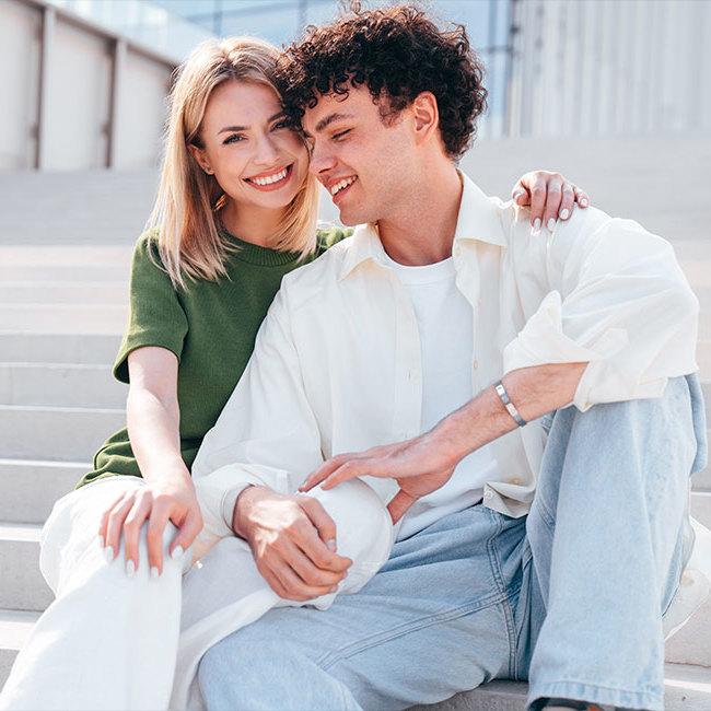 Et ungt par holder on hinanden på en trappe - hun smiler ind i kameraet mens han ser ned.